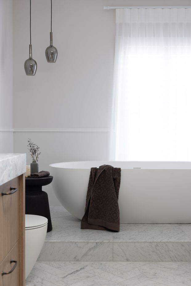 A bright, refined bathroom featuring a freestanding white oval bathtub set on a raised marble-tiled platform. The room has pale grey walls with white wainscoting, sheer white window curtains, two hanging glass pendant lights, a white toilet, a timber vanity with a pale stone benchtop, and a black stool or side table. The overall palette is soft white, grey, pale marble, timber, and dark brown accents.