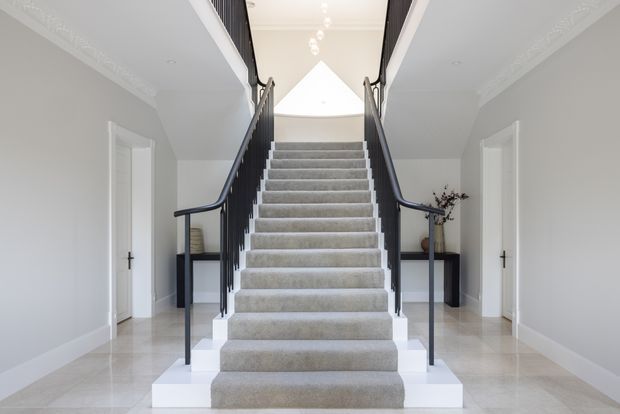 Bright, formal residential stairway with a broad central carpeted staircase, dark balustrades and handrails, white painted walls with ornate cornice detailing, polished pale stone flooring, and symmetrical doorways and console tables on either side. A skylight or roof lantern above the landing fills the space with natural light.