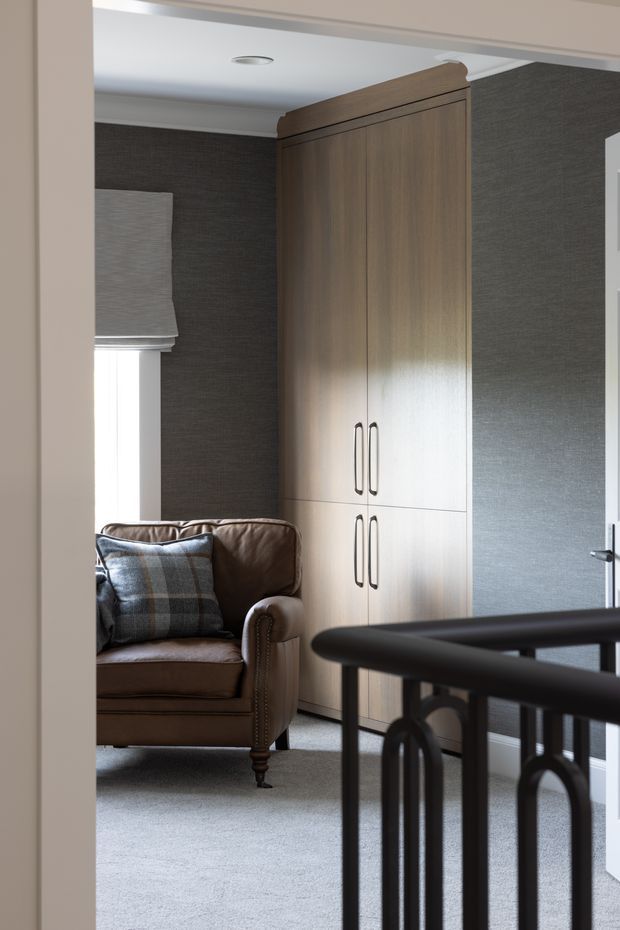 A refined sitting area viewed from a hallway or stair landing, featuring a brown leather armchair with a plaid cushion, a tall built-in timber storage cupboard, dark textured wallcovering, a pale carpet, and a black metal balustrade in the foreground. Soft daylight enters through a window with a Roman blind.