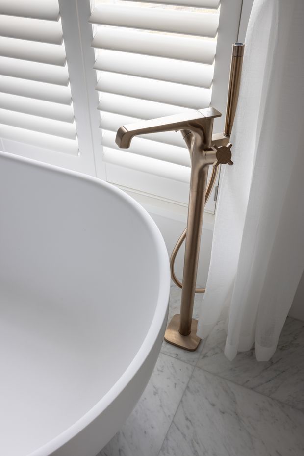 Close-up of a refined bathroom corner featuring a freestanding white oval bathtub, a bronze/brushed-gold floor-mounted tub filler with handheld shower, white plantation shutters, sheer white curtains, and pale veined marble flooring.
