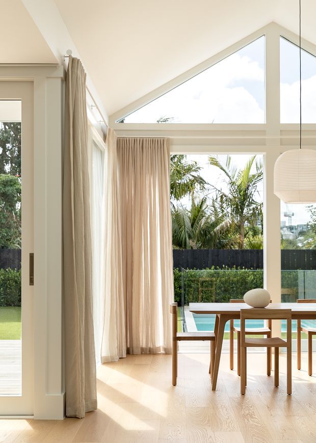 Bright, airy dining area with a warm neutral palette, pale timber flooring, full-height cream curtains, and large glazed openings including a distinctive triangular clerestory window. A timber dining table and chairs sit beside a pool, with tropical planting visible outside. A sculptural ribbed pendant hangs over the table.