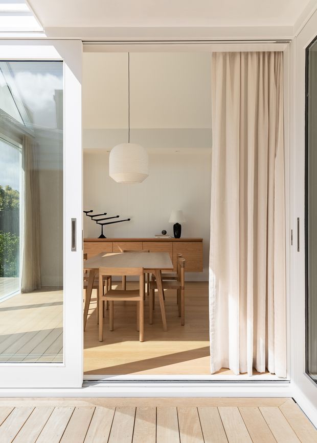 A bright, refined dining area viewed through large sliding glass doors. The space features a light timber dining table and chairs, built-in timber cabinetry along the rear wall, a white sculptural pendant light, full-height cream curtains, and abundant daylight. A timber deck and glazed indoor-outdoor threshold are visible in the foreground, with a sunlit adjacent space to the left.