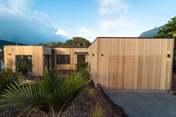 Single-storey contemporary residential building clad in warm vertical timber boards, composed of several simple box-like volumes around a recessed entry. A large timber-clad garage or entrance volume dominates the right side, with dark-framed windows and doors. Native-style landscaped planting and a concrete driveway sit in the foreground, while a steep metal roof is partly visible behind the facade.