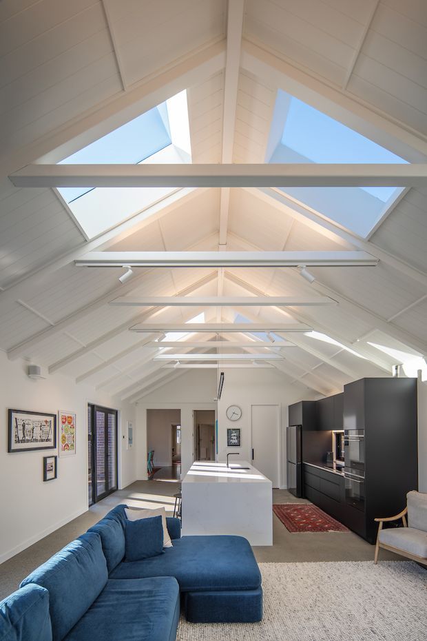 Bright open-plan interior with a large blue sectional sofa in the foreground, a white kitchen island, and matte black kitchen cabinetry along the right wall. The room has a white vaulted, timber-lined ceiling with multiple skylights and linear pendant or track lighting. Neutral carpet, framed wall art, dark-framed glazing, and a small dining or sitting area complete the space.