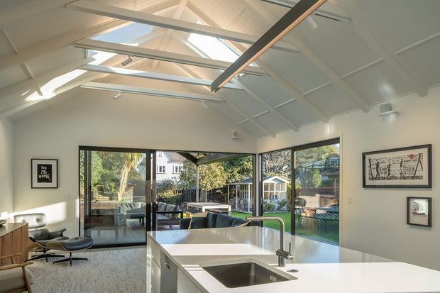 Bright contemporary open-plan kitchen and living area with a large white island, integrated stainless-steel sink and mixer tap, expansive glazed doors and windows, and a high vaulted ceiling with exposed painted structural beams and multiple skylights. The room opens toward a landscaped garden and outdoor entertaining area, with a lounge and framed artwork visible around the perimeter.