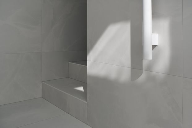Minimalist monochromatic bathroom interior with large light-grey tiled walls and floor, a built-in tiled step or bench, and a white cylindrical wall-mounted fixture. Strong directional sunlight creates geometric shadows across the smooth surfaces.