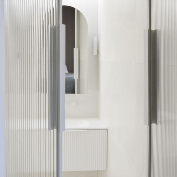 A compact, bright white bathroom vanity area viewed through vertically ribbed translucent glass doors or panels. The space features a white integrated benchtop and wall-mounted fluted vanity cabinetry, a tall arched mirror, white tiled walls, and minimalist vertical wall lighting. A partial reflection or view of a bedroom is visible through the mirror opening.