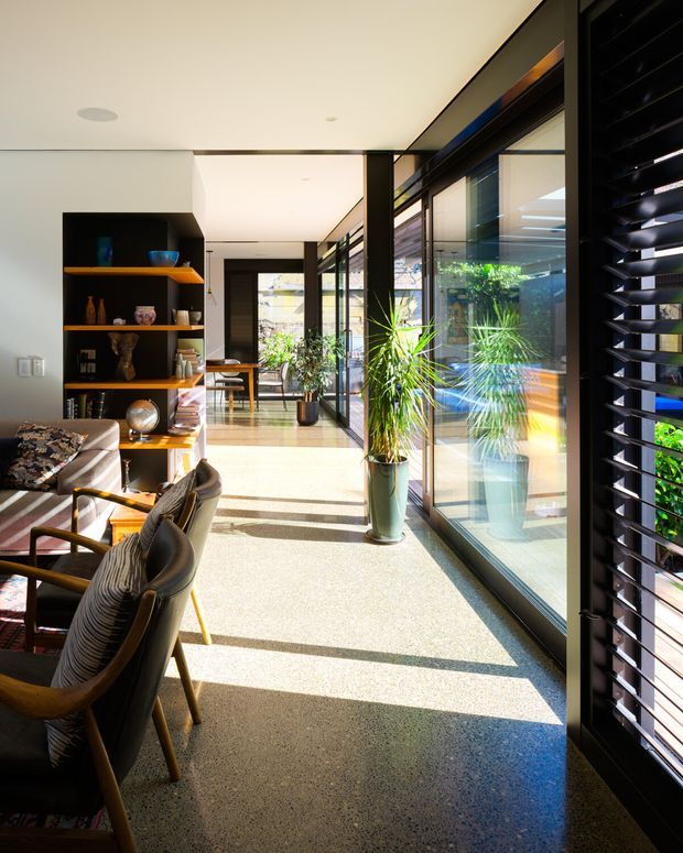 A bright contemporary interior corridor or living area with a polished exposed aggregate or terrazzo-like floor, full-height glazing, black-framed windows and doors, dark timber or painted shelving, and extensive indoor planting. Mid-century-inspired lounge chairs line the left side beside open display shelving, while strong sunlight casts long shadows across the floor. Black external-style louvres or blinds screen the glazing on the right.