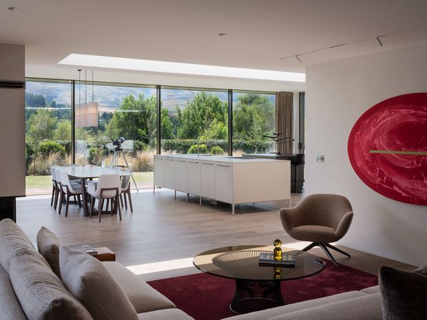 A refined open-plan living, dining and kitchen space with floor-to-ceiling glazing overlooking a landscaped garden and wooded hills. The room features a large pale sectional sofa, sculptural round coffee table, dining table with upholstered chairs, a minimalist white island and cabinetry, timber-look flooring, recessed ceiling lighting, and a bold glossy red circular wall artwork.