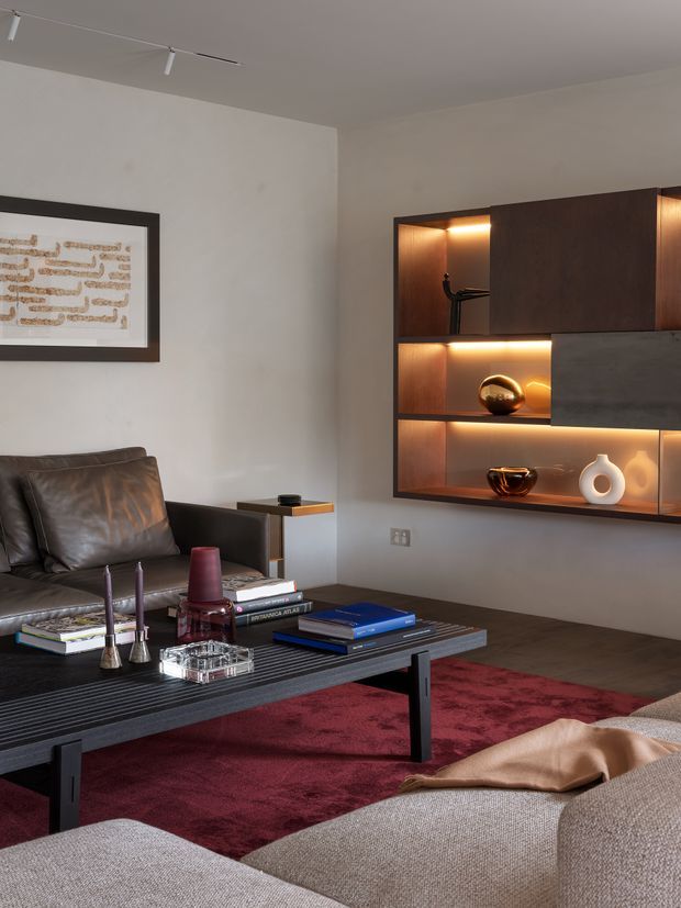 A refined living room with a dark leather sofa, large textured coffee table, burgundy rug, and a wall-mounted dark timber display cabinet with warm integrated lighting and decorative objects. Neutral plaster walls, track lighting, framed artwork, and a partially visible upholstered seating area create a warm, contemporary atmosphere.