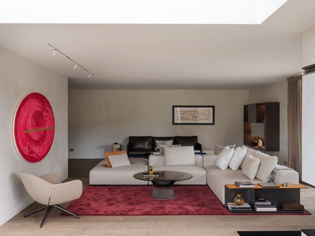 A refined contemporary living room with a large pale sectional sofa arranged around a low black oval coffee table, set over a deep red patterned rug. The room features light neutral walls and flooring, dark timber built-in shelving, floor-to-ceiling curtains, track lighting, a sculptural lounge chair, and a striking circular red wall artwork. A large ceiling opening or skylight brings in additional daylight.