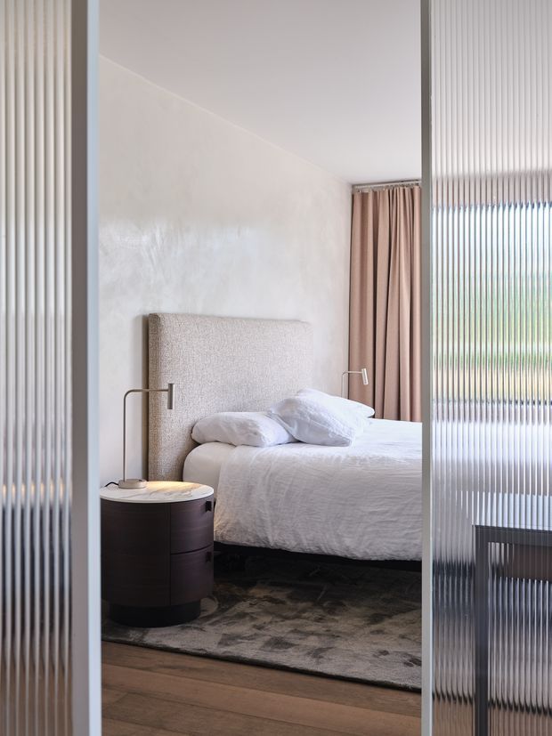 A refined contemporary bedroom viewed through vertically ribbed translucent glass doors. The room features a large upholstered headboard, white textured bedding, dark rounded bedside furniture, slim bedside lamps, taupe curtains, a large muted rug, timber flooring, and a softly textured pale wall. Natural light enters through the glazed opening on the right.
