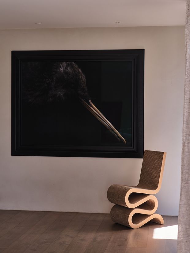 A quiet, warm-toned interior vignette with a large black-framed artwork featuring a feather against a dark background, a sculptural curved wood chair, pale plaster walls, timber flooring, recessed ceiling lights, and a sheer curtain at the right edge. Strong sunlight creates a bright patch on the floor, while the restrained furnishings give the space a refined, minimalist gallery-like atmosphere.