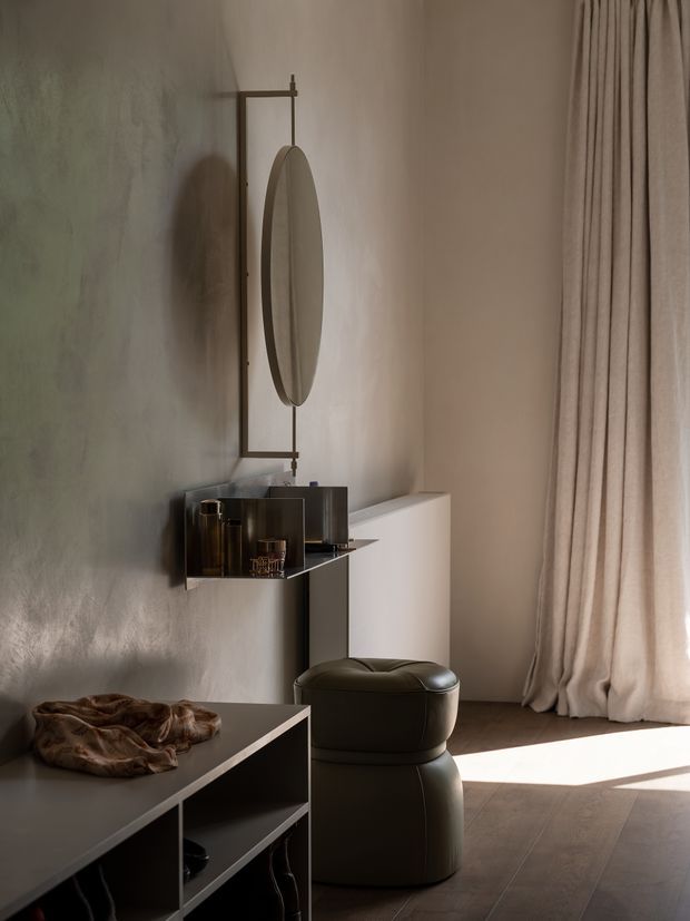 A softly lit contemporary interior vignette featuring a sculptural dark upholstered ottoman, a low open-shelving console, a wall-mounted oval mirror with a slim dark metal frame, and a compact drinks or bar niche. Textured warm grey plaster walls, full-height beige curtains, and dark timber flooring create a calm, refined atmosphere.