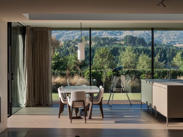 A contemporary open-plan dining and kitchen interior with full-height glazing overlooking a landscaped garden and mountainous rural scenery. The room features a pale dining table with upholstered chairs, timber flooring, sheer and heavy curtains, a streamlined island with integrated sink, and slim dark window frames. Natural light creates a calm connection between the interior and outdoor setting.