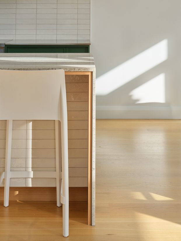 A sunlit minimalist kitchen or dining area detail featuring pale timber flooring, a compact island or counter with a light terrazzo-style benchtop, warm timber cabinetry, white tiled wall cladding, and a simple white stool. Strong geometric sunlight and shadows fall across the neutral wall and floor.