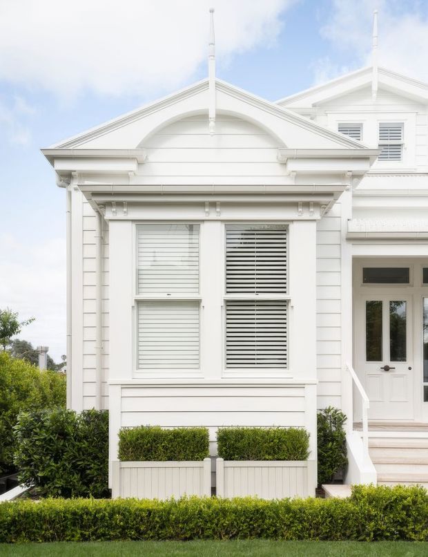 Bright exterior view of a refined white weatherboard house facade. The elevation features gabled roofs, layered classical trims, decorative brackets, horizontal cladding, plantation-shuttered windows, a white entry door with sidelights, and neatly clipped hedges and planter boxes.