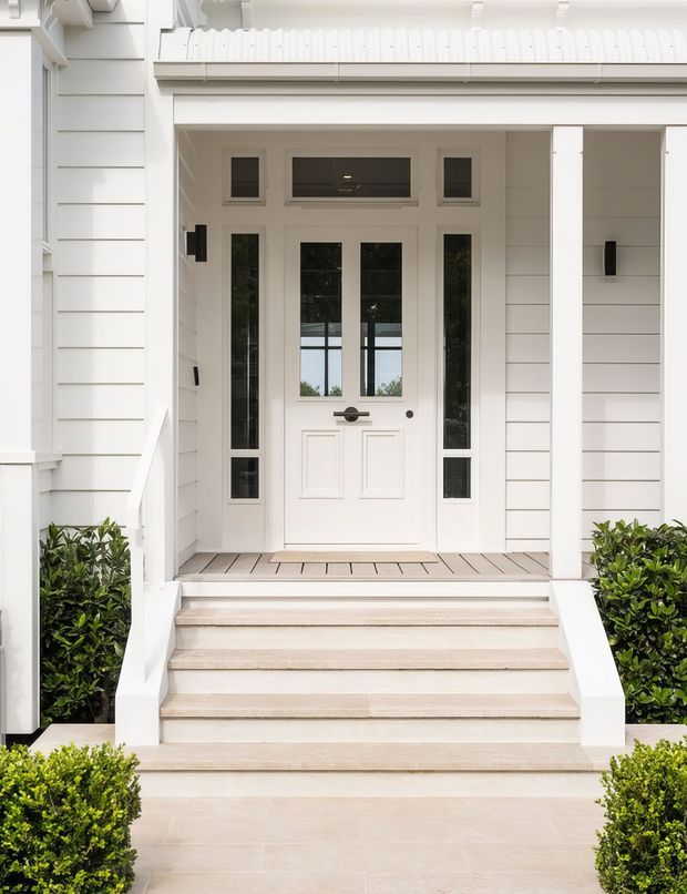 A symmetrical, close-up exterior view of a refined white residential entrance. A paneled front door with narrow glazed sidelights and transom sits beneath a covered porch, reached by broad pale stone steps. White horizontal weatherboard cladding, classical trim, dark hardware, timber porch decking, and neatly clipped green hedges create a clean, formal presentation.