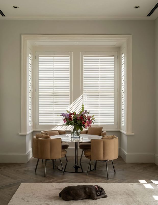 A refined dining nook set within a deep bay window, featuring white plantation shutters, layered wall and ceiling mouldings, and a round glass dining table surrounded by curved tan upholstered chairs. Herringbone timber flooring, soft neutral walls, a floral centrepiece, and a rug with a resting dog create a warm, elegant interior.