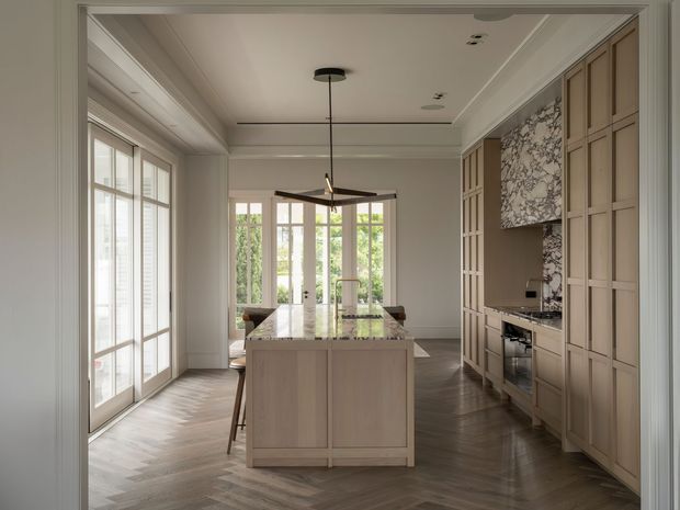 A refined, light-filled kitchen viewed through a wide doorway. The room features a large central island with a pale stone benchtop and integrated seating, full-height light timber cabinetry along the right wall, a patterned stone or marble backsplash and rangehood surround, dark herringbone timber flooring, tall glazed doors and windows, and a restrained coffered or stepped ceiling with a linear pendant light. The styling is elegant, warm, and contemporary-classic.