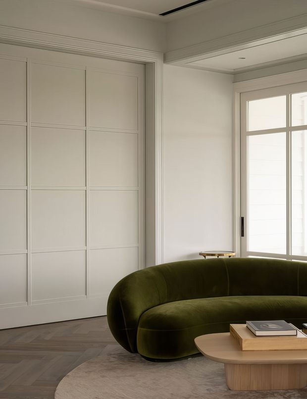 Refined living room with a curved olive-green upholstered sofa, rounded timber coffee table, pale textured rug, and dark herringbone timber flooring. The room features white painted paneled built-in cabinetry or doors, detailed traditional-style trim, tall divided-light windows, and a recessed ceiling detail. The restrained palette and sculptural furniture create a calm, contemporary yet classic interior.
