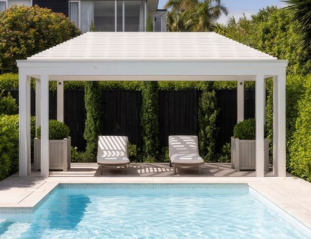 A bright outdoor pool area with a white architectural cabana or pergola positioned at the pool edge. The structure has a shallow pitched corrugated roof, square white posts, and a paved floor, with two reclining lounge chairs, planters, tall screening greenery, and a black slatted privacy fence behind it. A modern building and tropical landscaping are visible in the background.
