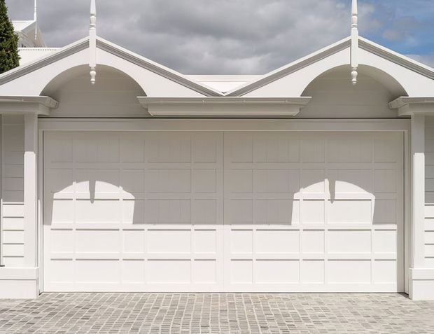 Front-facing exterior view of a symmetrical white garage facade with two large paneled garage doors, decorative arched porticos, gabled roof elements, ornamental finials, white weatherboard cladding, and a grey cobbled driveway under an overcast sky.