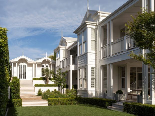 A formal residential courtyard or landscaped garden framed by elegant white multi-storey buildings. The architecture features symmetrical classical detailing, gabled roof forms, prominent cornices, louvred shutters, tall windows, covered balconies, white weatherboard-style cladding, and manicured hedges and lawn. A stepped walkway leads toward a pavilion-like structure at the rear under a bright blue sky.
