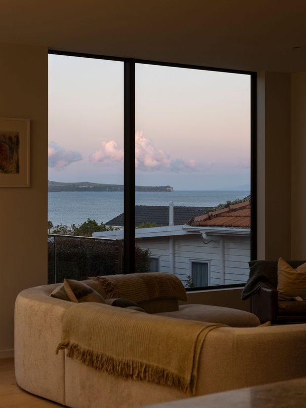 A warmly lit living room with a large black-framed picture window overlooking a calm coastal bay and nearby rooftops. A substantial beige sectional sofa with layered brown and taupe throws occupies the foreground, creating a cozy, relaxed interior. The room has a low, plain ceiling, warm neutral walls, timber flooring, and understated contemporary furnishings.