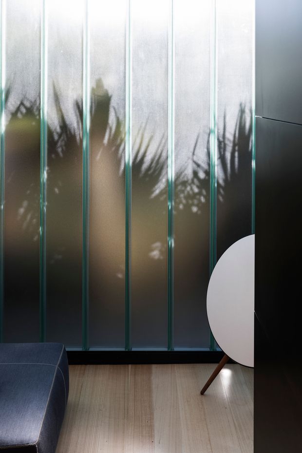 A moody contemporary interior corner featuring a tall wall of vertically divided translucent glass panels with dark framing, soft plant shadows, pale timber flooring, and dark built-in cabinetry. A partial navy upholstered sofa and a distinctive round white-fronted timber-legged piece of furniture are visible in the foreground.