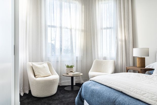 Bright, softly styled bedroom with a large corner of sheer-curtained windows, two upholstered boucle armchairs, a small round side table, bedside table and lamp, and a bed dressed in white bedding with a blue textured throw. The room has a calm, refined contemporary feel with dark carpet and neutral furnishings.