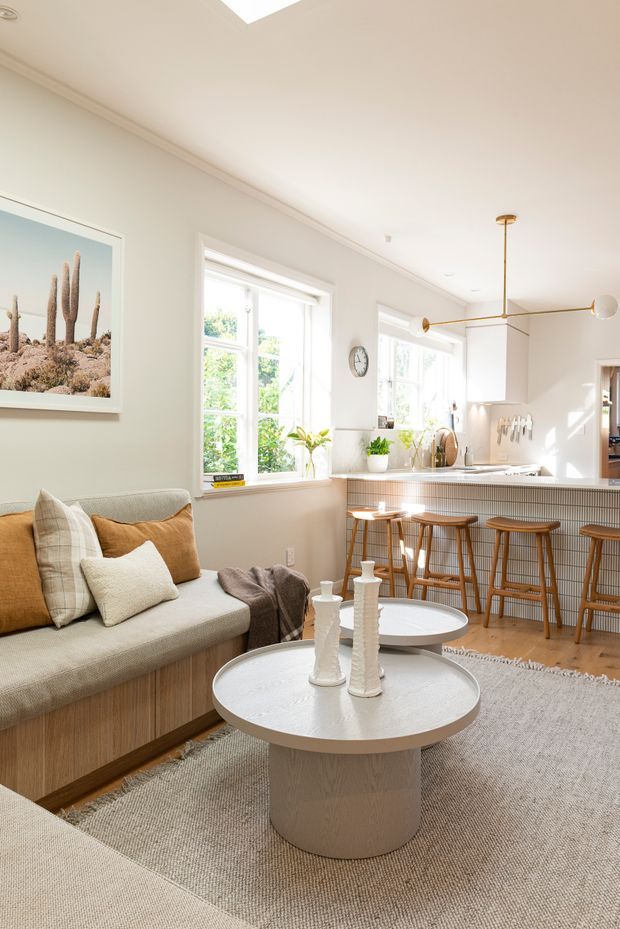 Bright open-plan living room and kitchen with a warm neutral palette. The room features a built-in upholstered sofa, round textured coffee tables, a large woven rug, white cabinetry, a tiled kitchen island with timber bar stools, and abundant natural light from large windows. Contemporary linear pendant lighting and desert-themed wall art add character.