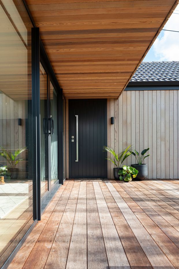A modern covered timber deck or entry terrace with a warm timber soffit and matching timber decking. Full-height black-framed glazing lines the left side, reflecting interior space, while a black vertical-panel front door and pale vertical timber cladding define the sheltered entry. Potted tropical plants add greenery near the doorway, and a tiled roof is visible beyond.