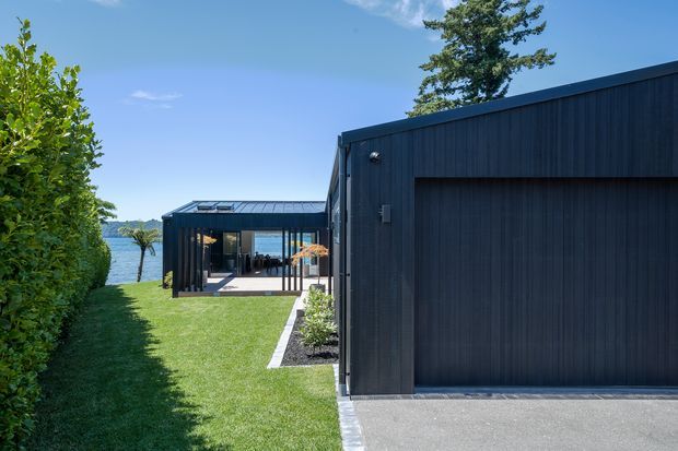 Exterior view of a contemporary dark-clad residential property beside a body of water. The foreground shows a large integrated garage with a flat roof and vertical dark cladding, while a glazed pavilion-like living area is visible beyond across a manicured lawn. The scene is sunny with clear blue skies and mature planting.