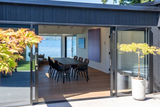 A contemporary dining area opens onto an outdoor deck through wide glazed sliding doors. The room features a dark timber dining table with black chairs, warm timber flooring, white walls, and a large vertical artwork panel. A blue waterfront view is visible beyond the glazing, with dark vertical exterior cladding and potted ornamental trees framing the entrance.