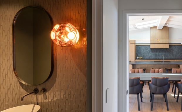A refined interior view looking from a compact bathroom into an adjoining dining and kitchen area. The bathroom features a tall rounded-rectangle mirror, wall-mounted basin tap, sculptural glowing wall light, and warm textured vertical-pattern wall tiles. Through the doorway, a contemporary dining table with upholstered blue chairs sits beneath timber detailing, with a dark kitchen splashback and cabinetry beyond.