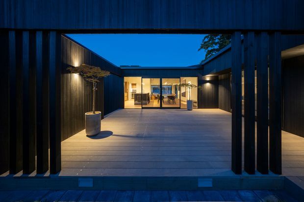 Twilight view through a dark timber-clad architectural opening into a sheltered courtyard deck. The courtyard has wide timber decking, sculptural potted trees, warm integrated wall lighting, and a glazed living or dining area at the far end. The composition emphasises strong geometric framing, a recessed courtyard layout, and contrast between the deep blue exterior and warmly lit interior.