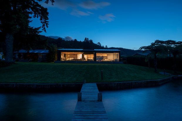 Twilight exterior view of a low, contemporary pavilion-style residence set behind a broad lawn and waterfront. The building has extensive warm-lit glazing, a flat or very low-pitched roof, and a simple horizontal form. A timber jetty projects across the water toward the property, with dark hills and mature trees forming the background.