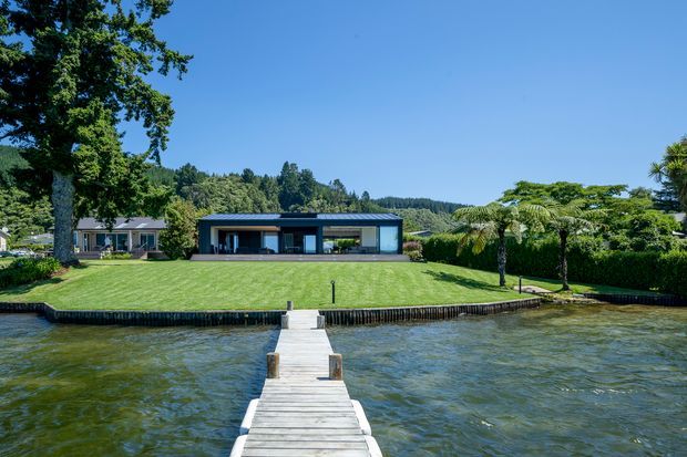Wide exterior view of a low, modern single-storey waterfront residence set behind a broad manicured lawn. The house has a dark horizontal facade, extensive floor-to-ceiling glazing, and a flat roof, with a timber jetty extending across the water toward the property. Mature trees, palms, hedges, and wooded hills form the landscaped backdrop.