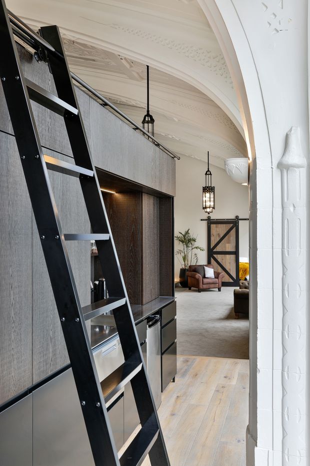 Interior view of a refined open-plan living area in a heritage-style building. A dark timber built-in joinery wall with integrated shelving and a black ladder occupies the foreground, opening toward a lounge with leather seating, plants, and a large barn-style timber door. Ornate white pressed ceiling details, curved cornices, arched openings, pendant lights, white painted brickwork, and pale timber flooring combine heritage architecture with contemporary industrial detailing.