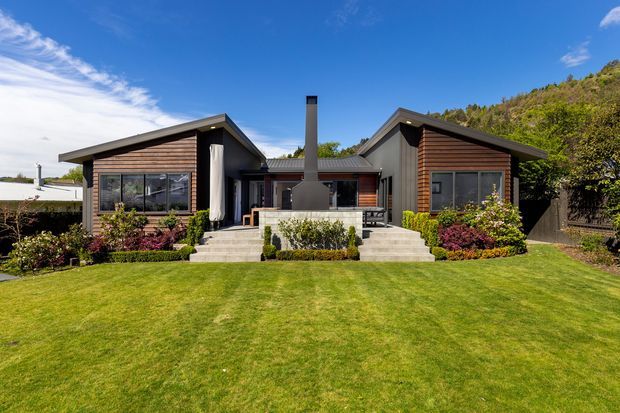 Front exterior view of a low-profile contemporary home with two dark timber-clad wings connected by a central entry and chimney volume. The house has sharply pitched asymmetrical gable roofs, extensive glazing, concrete steps, landscaped garden beds, and a broad lawn, set against a wooded hillside.