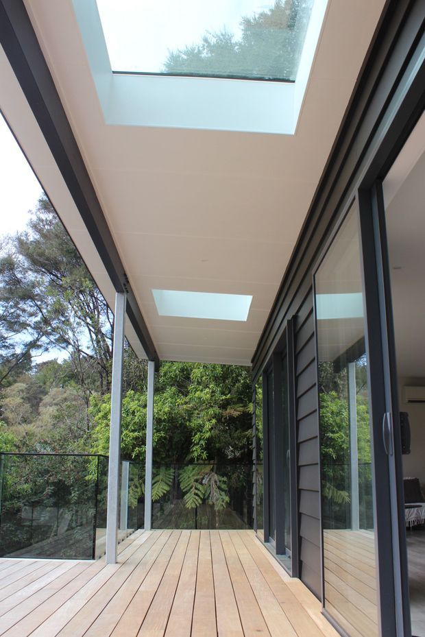A covered timber deck or terrace running alongside a modern glazed home, surrounded by dense greenery. Slim light-coloured structural posts support a white soffit with two rectangular skylights, while dark-framed floor-to-ceiling sliding glass doors and dark horizontal wall cladding line the building. Glass balustrading encloses the deck edge.