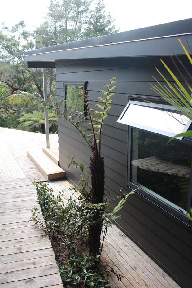Exterior view of a compact modern building set within dense vegetation, featuring dark horizontal cladding, a low-pitched roof with broad eaves, dark-framed windows, and a timber boardwalk/deck wrapping around the structure. The image emphasizes the facade, elevated walkway, and integration with the leafy landscape.