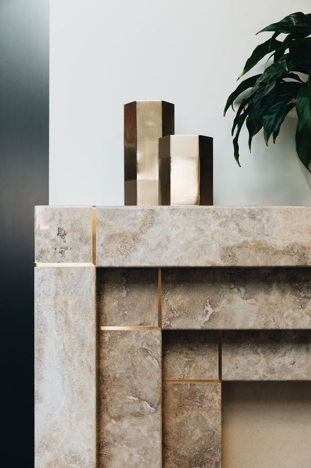 Close-up detail of a sculptural stone-clad fireplace or built-in feature wall. Large textured beige and taupe stone slabs form geometric surrounds and recessed shelving, with subtle brass-toned joints. Two art-deco-inspired rectangular wall lights or decorative objects sit on the mantel, against a pale wall with a leafy indoor plant at right.