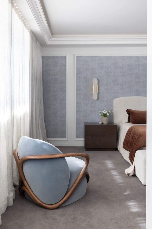 Elegant bedroom interior with a soft, neutral palette. The room features grey-blue textured wall panels framed with white moulding, a pale upholstered bed with brown bedding, a dark timber bedside table, a sculptural timber-framed lounge chair, sheer full-height curtains, and detailed white ceiling cornices. Natural light creates a calm, refined atmosphere.