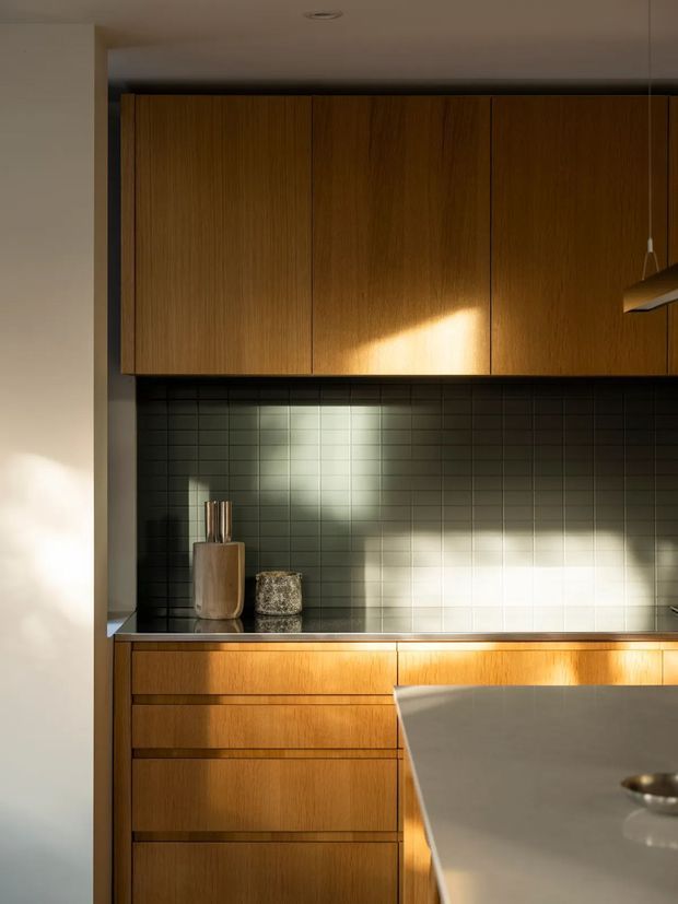 A close-up view of a warm, contemporary kitchen featuring vertically grained timber cabinetry, a dark green-grey square tile splashback, and a light stone benchtop or island in the foreground. Strong late-afternoon sunlight creates dramatic highlights and shadows across the cabinetry and tiled wall. Minimal countertop accessories and a modern pendant light are visible.