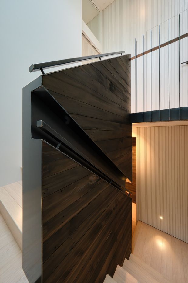 Interior architectural photograph looking up and along a sculptural staircase. Dark timber-clad stair stringers and stair soffits contrast with pale timber flooring and vertically ribbed light wall panels. Slim dark metal handrails and balustrade lines emphasize the dramatic geometry, while warm concealed lighting glows beneath the upper landing. The space appears tall and open, with a refined contemporary architectural character.