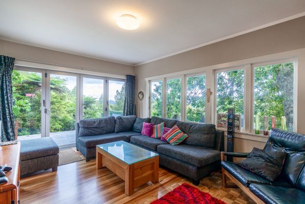 Bright, welcoming living room with large white-framed windows and glazed doors opening to a leafy outdoor setting. The room features warm timber parquet flooring, taupe walls with simple white trim, a large dark grey sectional sofa with colourful striped cushions, a timber coffee table with a glass top, a matching ottoman, a black leather armchair, and patterned curtains. Natural light and greenery create an airy connection to the outdoors.