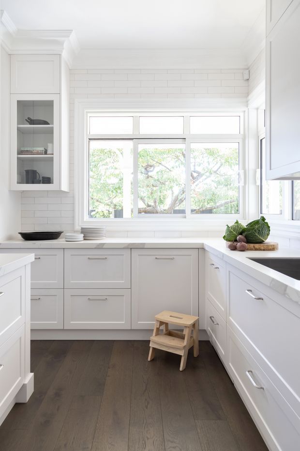 Bright, compact white kitchen with U-shaped cabinetry, shaker-style paneled doors, white subway-tile walls, pale benchtops, dark timber flooring, and a large multi-pane window providing abundant natural light. Open shelving displays dishes and cookware, while fresh produce sits on a timber board near the sink.