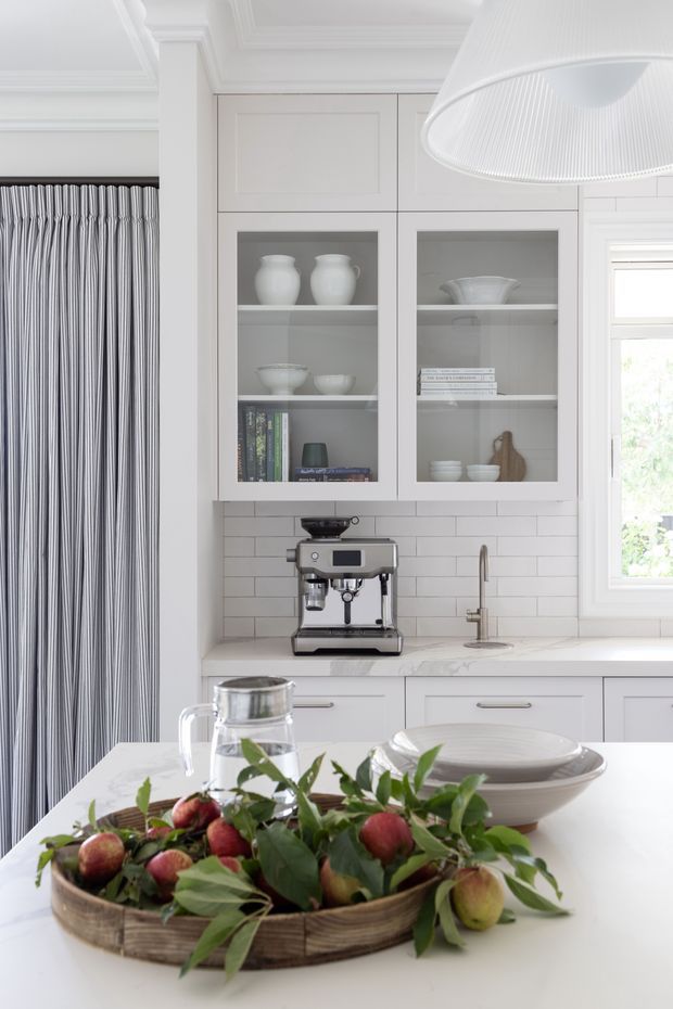 Bright, refined kitchen interior featuring white built-in glass-front cabinetry, a white subway-tile backsplash, pale stone benchtops, a compact coffee station, and a farmhouse-style sink beneath a window. A large white pendant light, striped curtains, and a dining island with neutral tableware and a fruit arrangement complete the scene.
